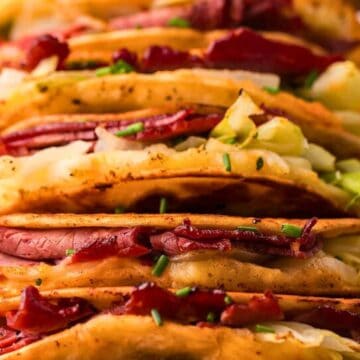 Four crispy tacos filled with sliced meat, cabbage, and chopped chives on a white rectangular plate.