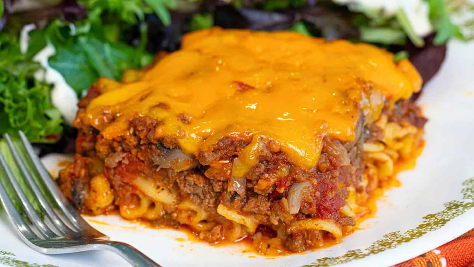 A slice of cheesy baked lasagna with ground meat and sauce, served with a side of mixed greens on a plate.