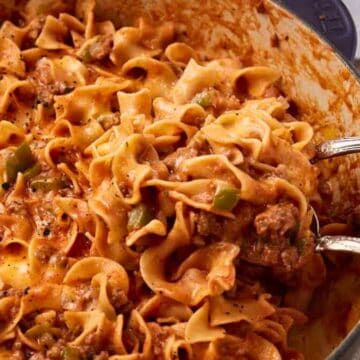 A pot of cheesy beef noodle casserole with two serving spoons, showing melted cheese and ground beef.