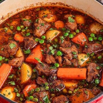 A hearty beef stew with carrots, potatoes, and peas simmering in a red Dutch oven on a wooden table.