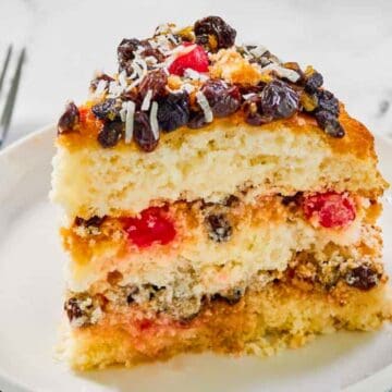 A slice of layered fruitcake with raisins and cherries on a white plate, fork on the side.