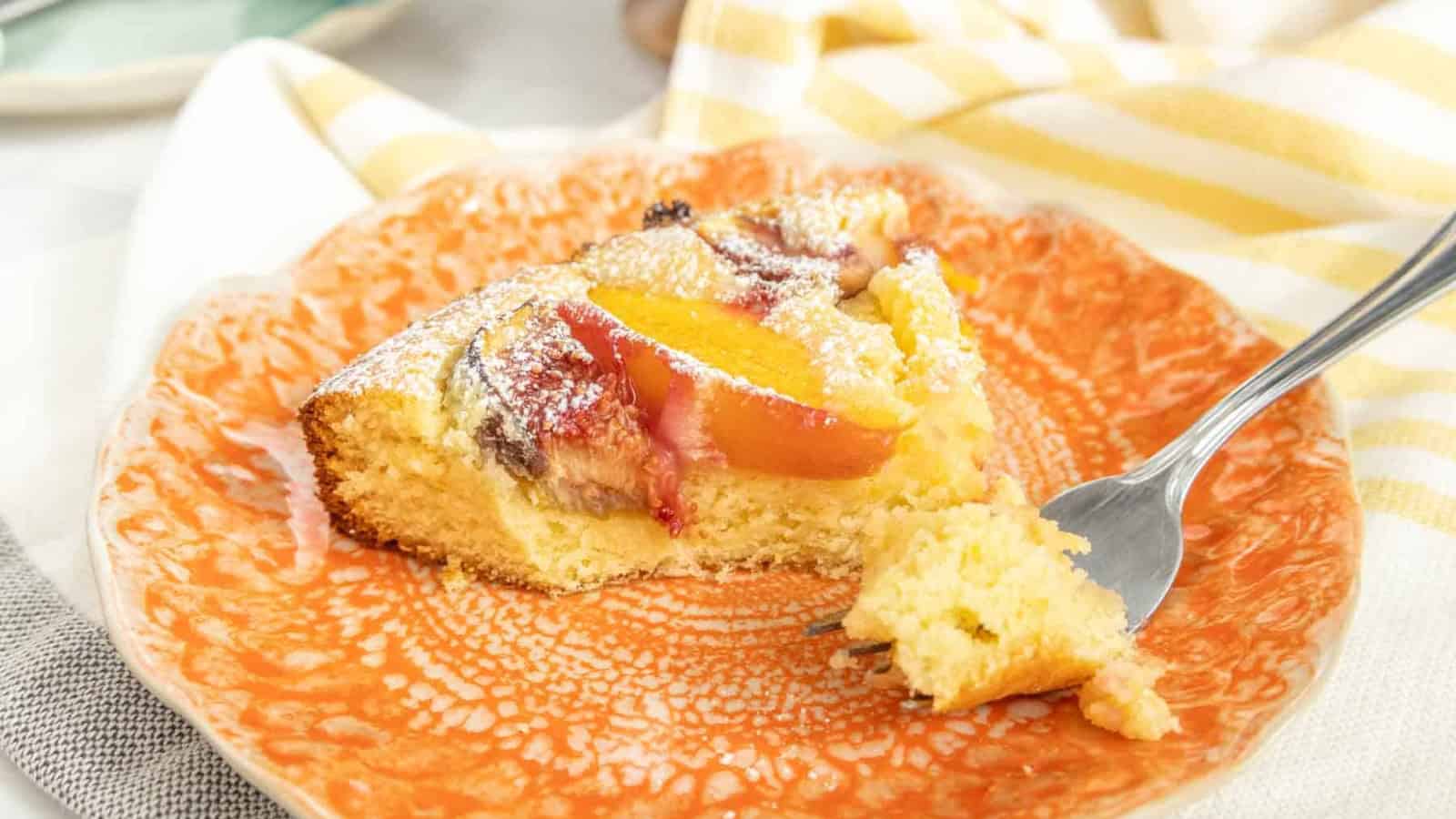 A slice of fruit-topped cake with powdered sugar on an orange plate, with a fork beside it.