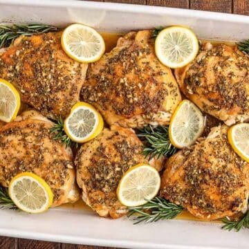 Baked chicken thighs with lemon slices and rosemary in a white baking dish on a wooden table.