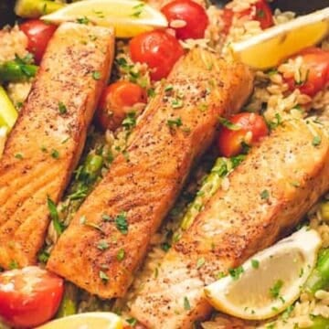 Three cooked salmon fillets on a bed of rice with cherry tomatoes, asparagus, and lemon wedges in a skillet.