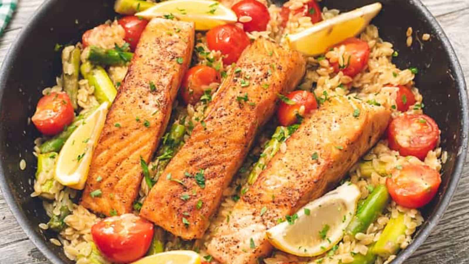 Three cooked salmon fillets on a bed of rice with cherry tomatoes, asparagus, and lemon wedges in a skillet.