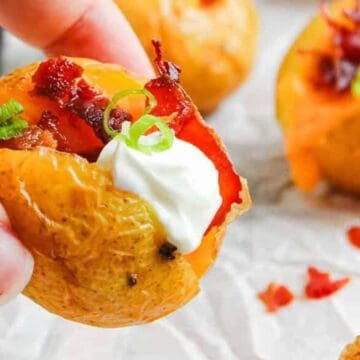A hand holds a mini baked potato topped with sour cream, bacon bits, cheese, and sliced green onions.