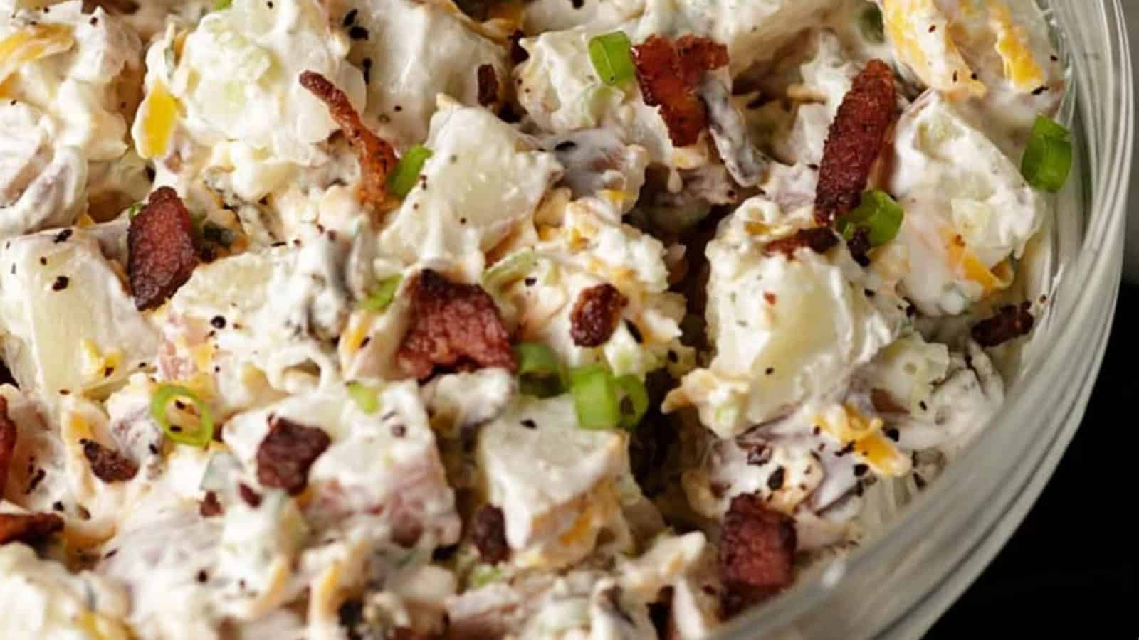 A close-up of a bowl of creamy potato salad with chopped green onions, crispy bacon pieces, and black pepper on top.