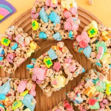 Colorful marshmallow cereal bars arranged on a wooden tray, with rainbow paper decorations on a yellow background.