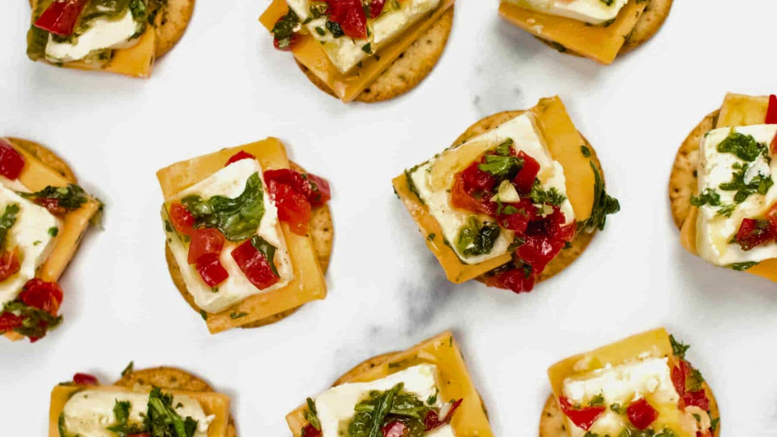 Round crackers topped with cheddar, brie, diced red peppers, and chopped herbs on a white surface.