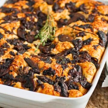 Baked casserole topped with mushrooms and herbs in a white dish beside plates, forks, and a knife.