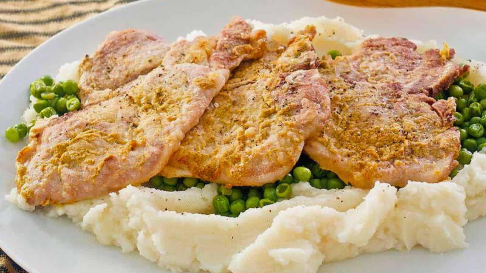 Three pieces of breaded pork on mashed potatoes with green peas on a white plate.