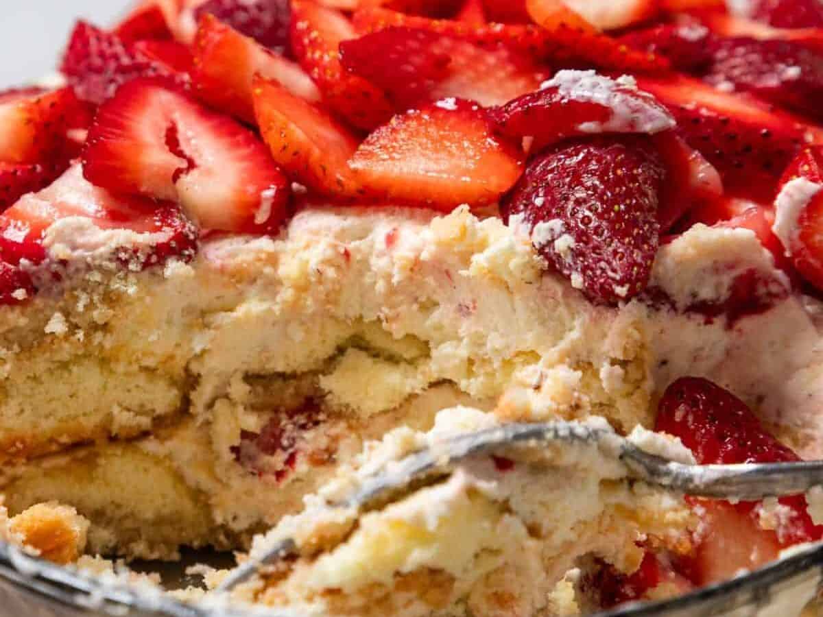 A close-up of a No-Bake Strawberry Tiramisu shows layers of cream, sponge cake, and sliced strawberries on top. A serving has been scooped out, revealing the dessert's delicious texture inside a glass dish.
