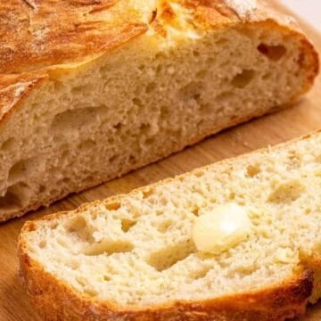 A sliced loaf of rustic bread with a pat of butter on one slice, on a wooden cutting board.