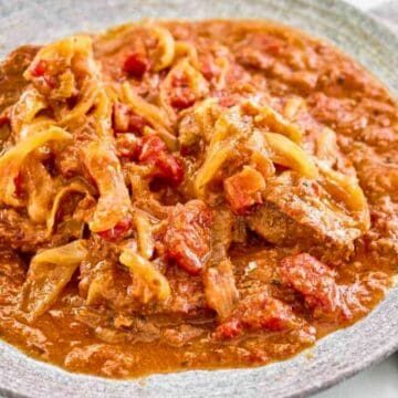 A plate of beef stew with onions and tomatoes in a rich, reddish-brown sauce.