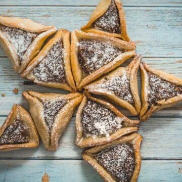 Hamantaschen cookies, a classic treat from Purim recipes, are arranged in a star shape on a rustic blue wooden surface and dusted with powdered sugar.