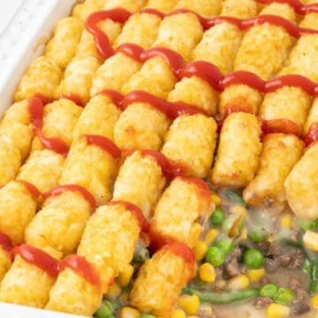 A casserole topped with tater tots and ketchup, filled with mixed vegetables and ground beef.