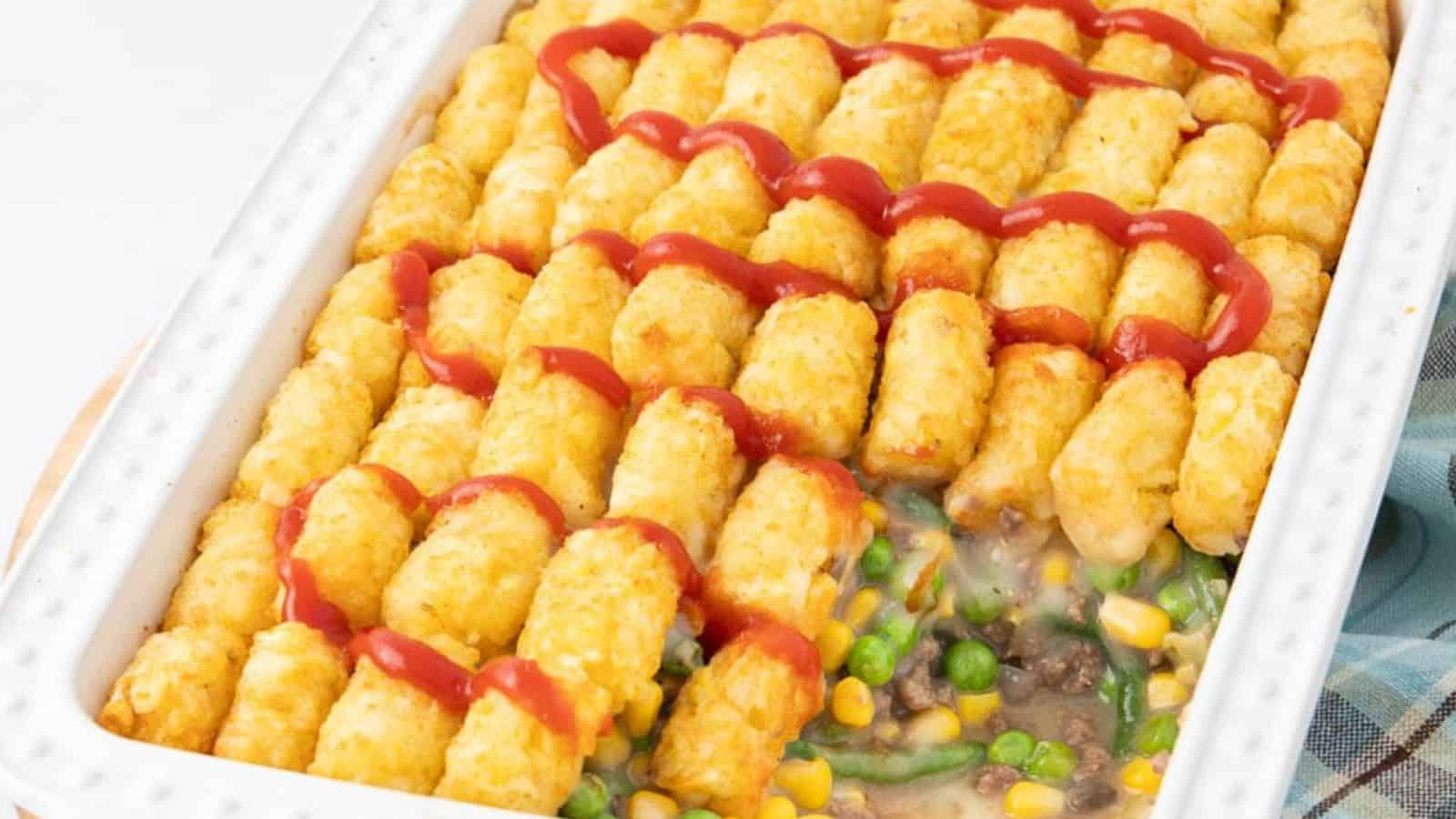 A casserole topped with tater tots and ketchup, filled with mixed vegetables and ground beef.