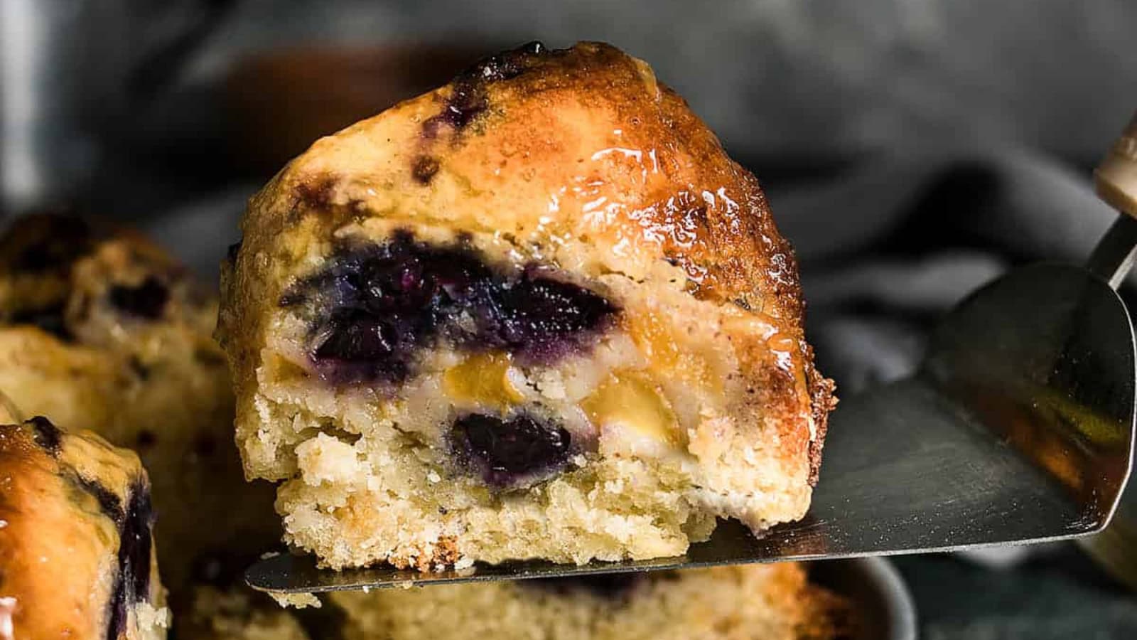 A close-up of a blueberry muffin slice with visible blueberries and a moist, golden crumb on a spatula.