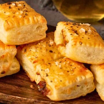 Golden, flaky pastry squares topped with salt and pepper, stacked on a wooden serving board.