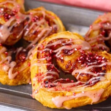 Heart-shaped pastries with pink icing and red sprinkles on a metal tray.