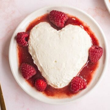 A heart-shaped dessert with raspberries and sauce on a white plate, next to a gold spoon.