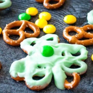 Pretzels dipped in green icing with green and yellow candies and caramel chocolates on a dark surface.