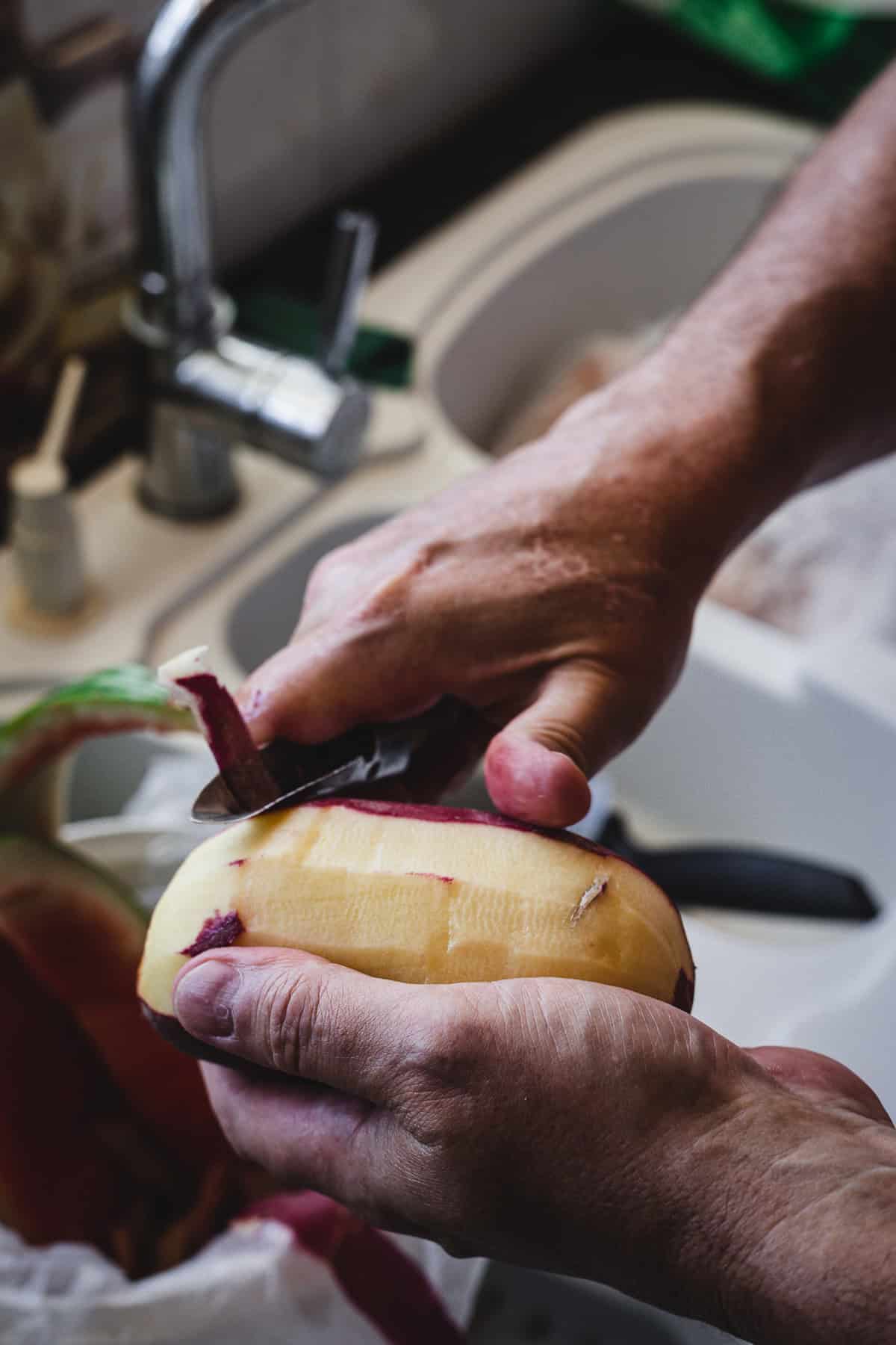 Hands peeling a potato with a knife over a kitchen sink, water faucet and peels visible in the background—prepping potatoes for classic Russian Fried Potatoes.