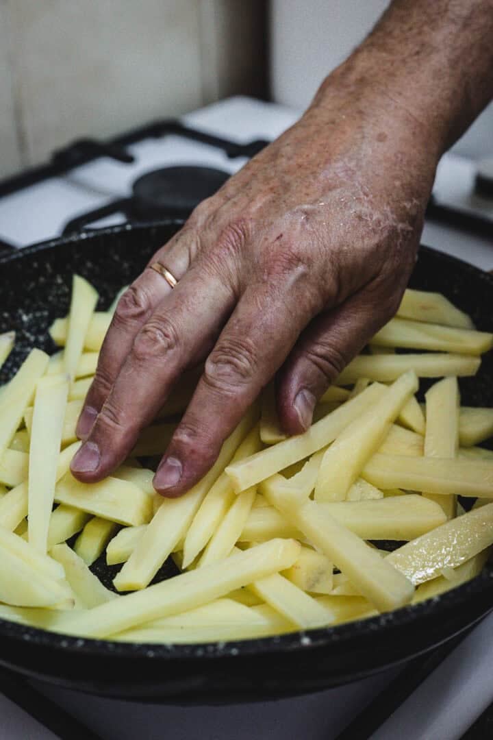 A hand arranging raw potato fries in a frying pan on a stove, preparing classic Russian Fried Potatoes.