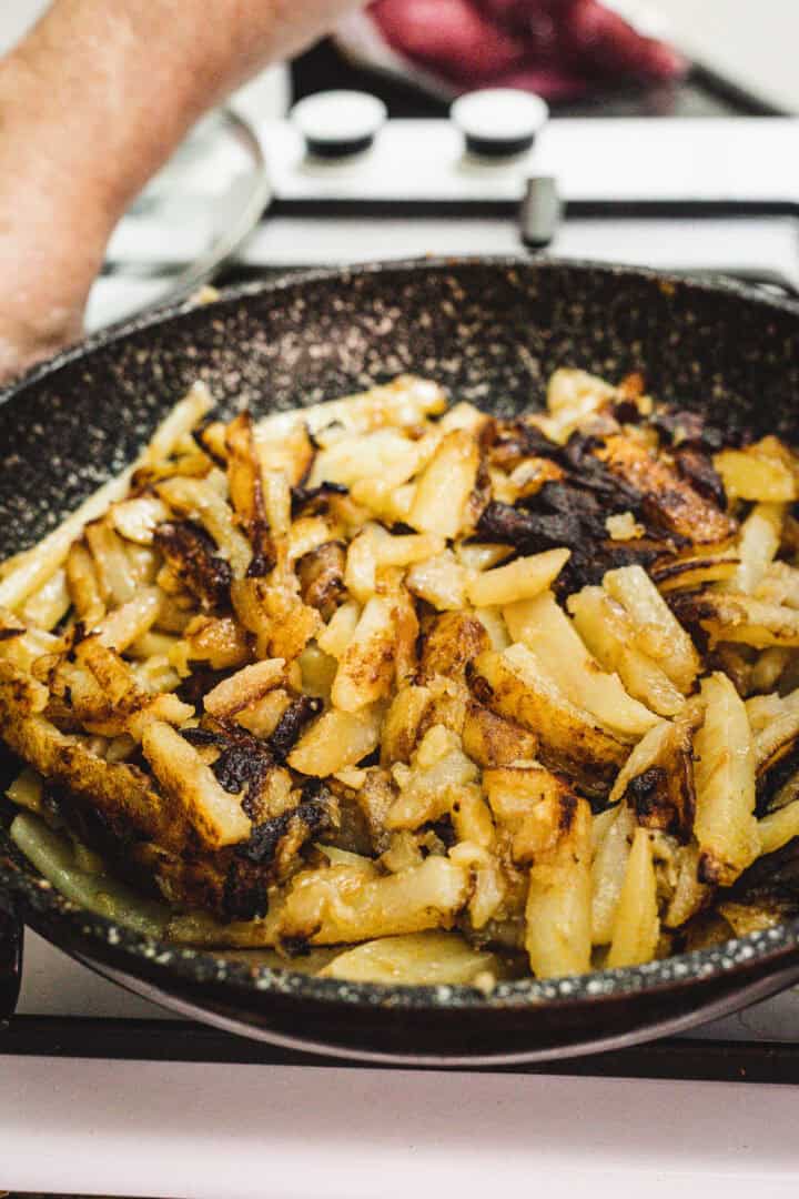 Russian Fried Potatoes sizzle in a skillet on the stove, with a hand visible in the background.