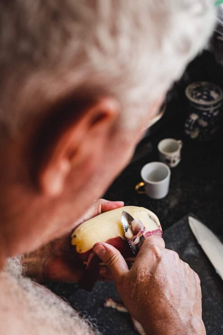 An elderly person peels a potato with a spoon at a kitchen counter, preparing ingredients for Russian Fried Potatoes next to a knife and cups.