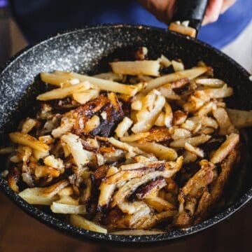 A hand holds a pan filled with crispy, golden-brown fried potatoes.
