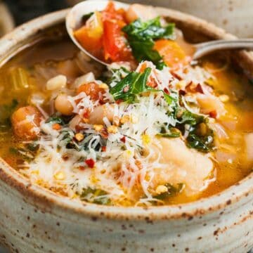 A bowl of vegetable soup topped with grated cheese and herbs, with a spoon resting on the bowl’s edge.