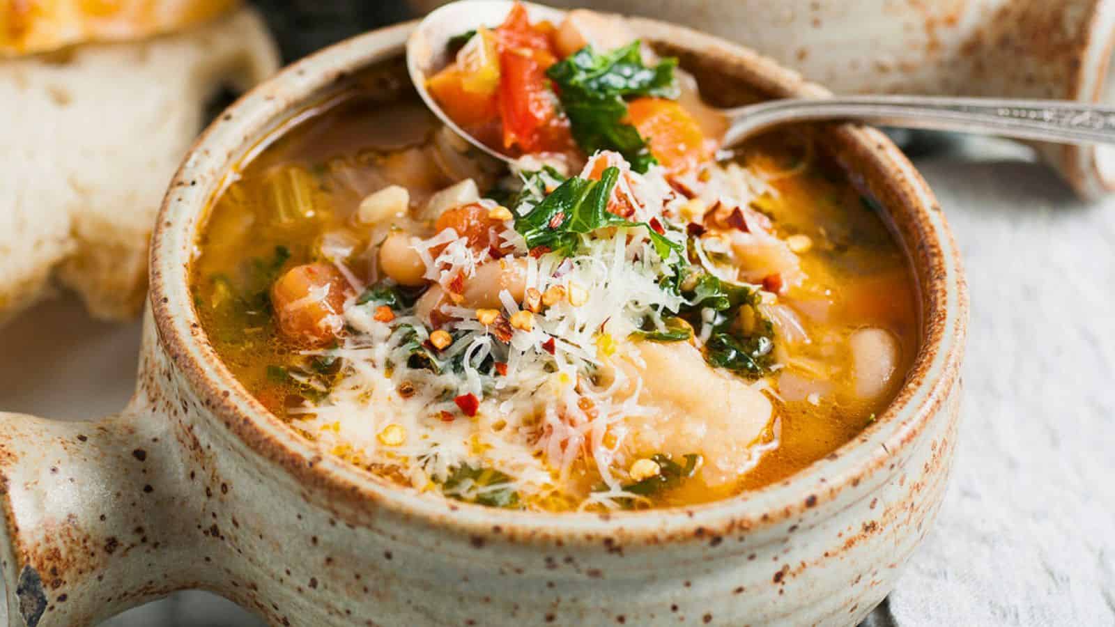 A bowl of vegetable soup topped with grated cheese and herbs, with a spoon resting on the bowl’s edge.