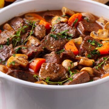 A white pot filled with beef stew, carrots, mushrooms, and herbs on a table.