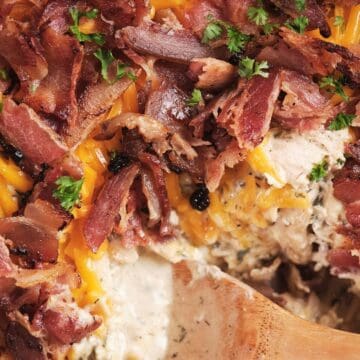 Closeup of creamy chicken casserole topped with melted cheese, crispy bacon, and chopped parsley, with a wooden spoon.