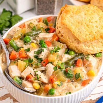A bowl of creamy chicken and vegetable stew topped with a biscuit, with parsley and a blue checkered napkin nearby.