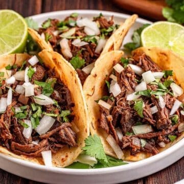 Three beef tacos topped with chopped onions and cilantro, served with lime halves on a white plate.