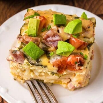 A slice of breakfast casserole topped with avocado and tomato on a white plate with a fork.