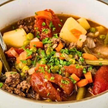 A bowl of beef stew with potatoes, carrots, green beans, peas, and tomatoes, garnished with fresh herbs.
