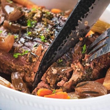 A fork and knife slicing pot roast with carrots, onions, and celery in a white baking dish.