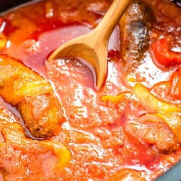 Sausages simmering in a rich tomato sauce with onions, stirred by a wooden spoon.