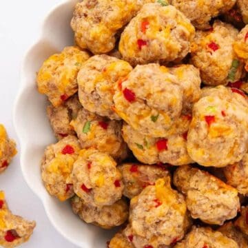 Cheesy sausage balls with bits of red and green pepper in a white bowl, with a few on the side.