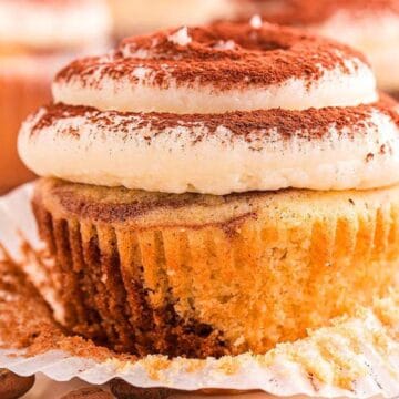A tiramisu cupcake with creamy frosting and cocoa powder, partially unwrapped with a bite taken out.