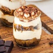 A glass jar filled with layered tiramisu, topped with cocoa powder and chocolate shavings, on a wooden board.