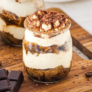 A glass jar filled with layered tiramisu, topped with cocoa powder and chocolate shavings, on a wooden board.