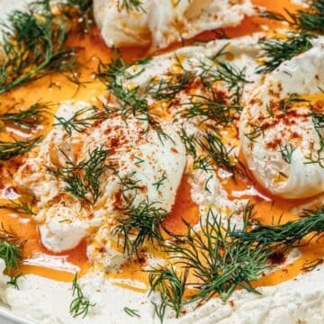 Creamy yogurt topped with poached eggs, paprika oil, and fresh dill on a white plate.