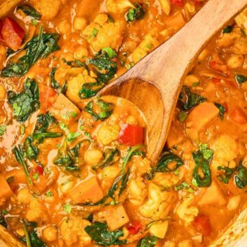 A pot of creamy vegetable curry with chickpeas, spinach, and a wooden spoon stirring the dish.