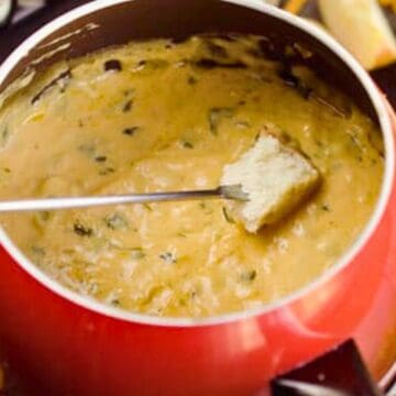 A piece of bread is dipped into a pot of melted cheese fondue with herbs.