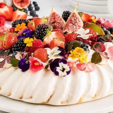 A pavlova, perfect for spring desserts, topped with whipped cream, fresh berries, figs, and colorful edible flowers on a white plate.