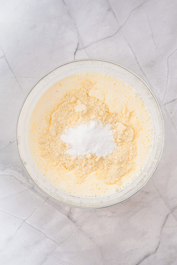 A glass bowl with flour, baking powder, and wet batter on a marble surface, ready to be mixed.